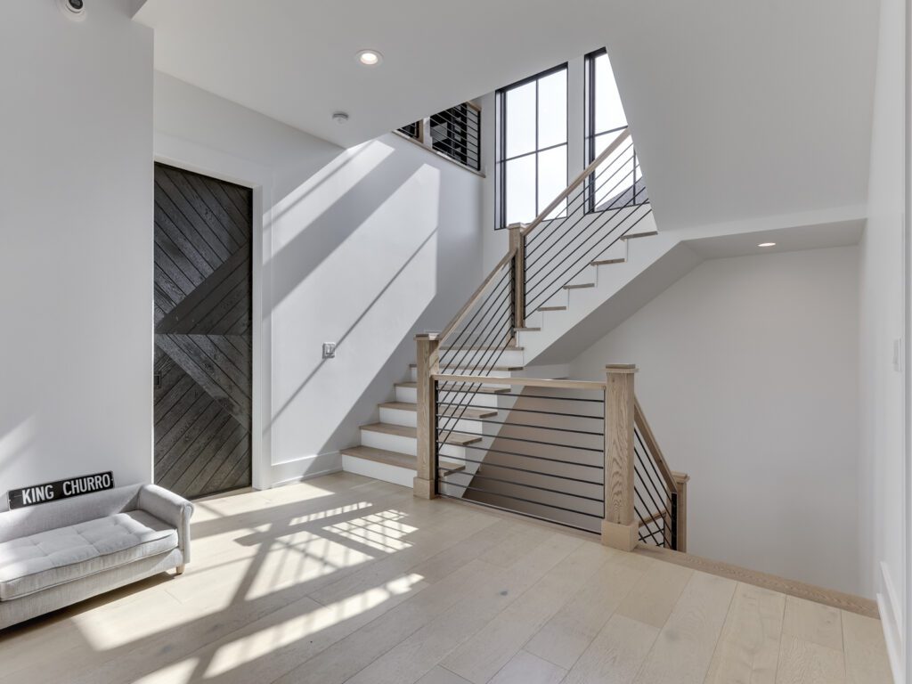 Bright and modern entryway featuring a staircase, sleek railing, and a geometric door. Sunlight casts shadows on the light hardwood floor.